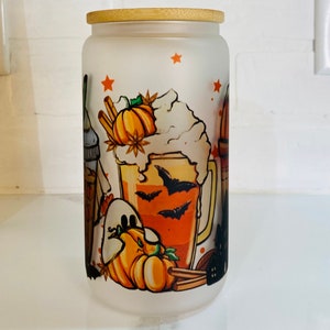 May include: A frosted glass jar with a bamboo lid features a Halloween-themed design. The design includes a pumpkin spice latte with whipped cream, bats, and a ghost. The jar is perfect for storing treats or other small items.