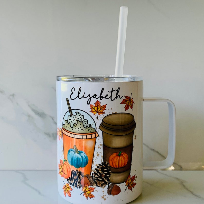 May include: White stainless steel tumbler with a handle and a clear lid. The tumbler features a fall-themed design with two coffee cups, one with whipped cream and the other with a brown lid. The design also includes autumn leaves, pumpkins, and pine cones. The tumbler has the name "Elizabeth" printed on the front.
