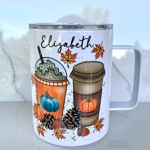May include: White stainless steel travel mug with a black and brown illustration of a pumpkin spice latte and a pumpkin iced coffee. The mug has a black handle and the name "Elizabeth" is printed on the front.