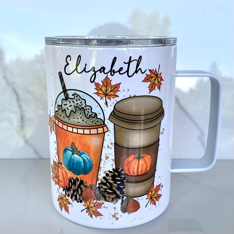 May include: White stainless steel travel mug with a black and brown illustration of a pumpkin spice latte and a pumpkin iced coffee. The mug has a black handle and the name "Elizabeth" is printed on the front.