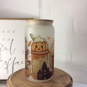 May include: A frosted glass tumbler with a bamboo lid and a clear straw. The tumbler features a fall-themed design with a pumpkin-shaped coffee cup, a ghost, a house, and leaves. The tumbler is sitting on a wooden stand.