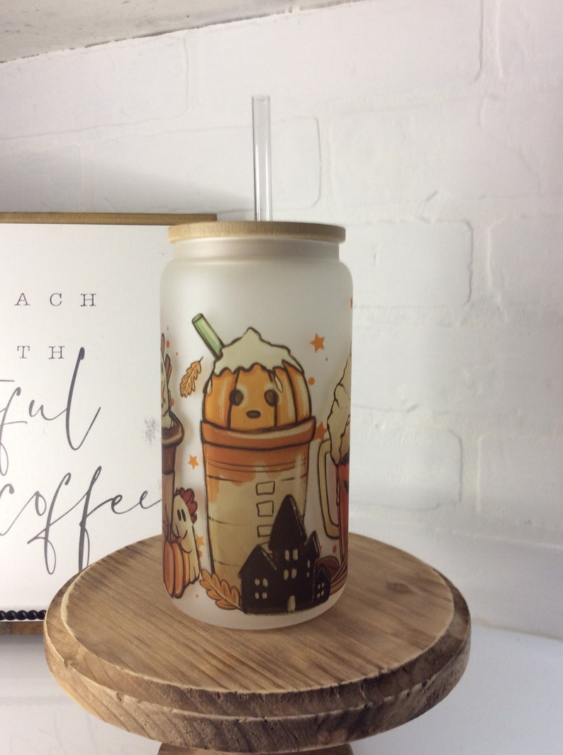 May include: A frosted glass tumbler with a bamboo lid and a clear straw. The tumbler features a fall-themed design with a pumpkin-shaped coffee cup, a ghost, a house, and leaves. The tumbler is sitting on a wooden stand.
