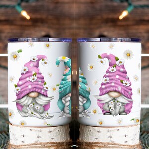 May include: Two white ceramic mugs with a pink and blue gnome design. The gnomes are wearing hats with white flowers and have white beards. The mugs are surrounded by white daisies.