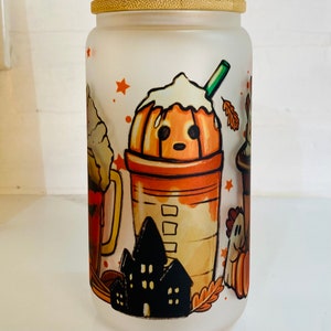 May include: A white frosted glass jar with a bamboo lid features a Halloween-themed design. The design includes a pumpkin spice latte with whipped cream, a ghost, a house, and a fall leaf.