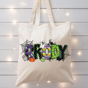 May include: A cream-colored tote bag with the name "Brady" in a Halloween-themed design. The letters are in purple, green, and orange with spiderwebs, a ghost, and a pumpkin. The bag has two handles.
