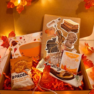 May include: A gift box containing autumn-themed treats. It includes a book with stickers, a packet of oat crumbles, a chocolate bar, and a tea bag. The box is filled with orange paper and artificial autumn leaves.