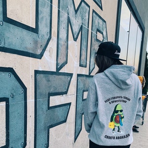 Può includere: Una persona indossa una felpa con cappuccio grigia con un berretto da baseball nero che dice "The Jump". La felpa ha una grafica di un avocado con un viso e il testo "Буду говорить только в присутствии своего авокадо" sul retro.