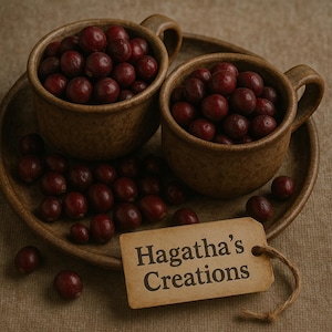 Könnte beinhalten: Zwei braune Keramikbecher, gefüllt mit frischen, dunkelroten Cranberries, stehen auf einem passenden Teller. Verstreute Cranberries umgeben die Becher und den Teller. Ein kleines Etikett mit dem Text "Hagatha's Creations" hängt am Teller.