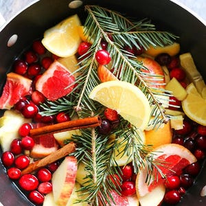 Puede incluir: Una olla negra llena de agua, cítricos, arándanos, rodajas de manzana, palitos de canela y ramitas de hoja perenne. La disposición incluye rodajas de pomelo, limón y manzana, con arándanos rojos brillantes.