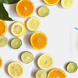May include: Overhead shot of sliced oranges, lemons, and limes scattered on a white surface. Green leaves and a glass of juice with a blue straw are also present, creating a refreshing, summery theme.
