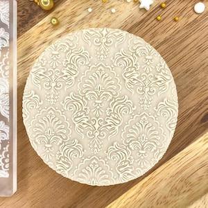 May include: A white silicone rolling pin with a damask pattern. The rolling pin is on a wooden surface with gold and white decorative accents.