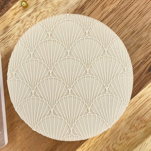 Könnte beinhalten: Ein runder, cremefarbener Keksstempel mit einem fächerförmigen Art-Deco-Design. Daneben befindet sich eine transparente Acrylschablone mit dem gleichen Design. Beide liegen auf einer Holzoberfläche.