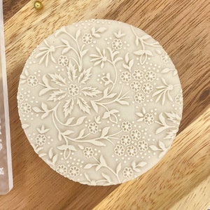 Könnte beinhalten: Ein weißer Silikon-Kuchen-Dekor-Stempel mit einem floralen Design. Der Stempel ist rund und hat ein erhabenes Design aus Blumen und Blättern.