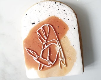 May include: A white sugar cookie decorated with a line drawing of a person wearing a headscarf. The background is a light brown and orange watercolor wash with black speckles.