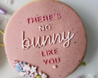 May include: Pink iced sugar cookie with white sprinkles. The cookie has pink icing with the text "There's no bunny like you" written in white and pink icing.
