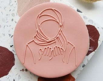 May include: A pink cookie with a white outline of a person wearing a headscarf. The cookie is on a terrazzo surface.