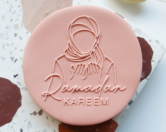 May include: A pink round stamp with a line drawing of a person wearing a headscarf. The text "Ramadan Kareem" is written in cursive below the drawing.
