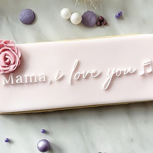May include: A pink rectangular cookie decorated with white icing. The icing says "Mama, I love you" with a musical note. A pink sugar flower is on the left side of the cookie.