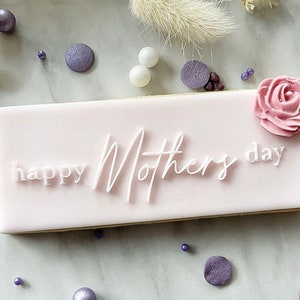 May include: A pink rectangular cookie with white icing and the text "happy Mother's day" on top. A pink sugar flower is on the right side of the cookie.