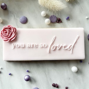 May include: A pink rectangular cookie with white icing and the words "you are so loved" written on it. A pink sugar rose is on the top left corner of the cookie.