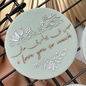 Puede incluir: Una galleta redonda de color verde claro con diseños florales blancos y dorados y el texto "I love you so much" en letras doradas.