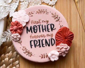 May include: Pink round cake with the text "First my Mother forever my Friend" in black lettering. The cake is decorated with pink and red flowers.