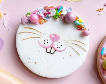May include: A white sugar cookie decorated with a bunny face and pink and gold accents. The cookie is topped with colorful sprinkles.