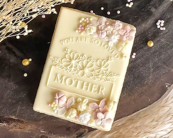 May include: White chocolate cookie decorated with pink and yellow flowers and the text "You are so loved Mother".