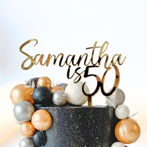 May include: Gold cake topper with the text "Samantha is 50" on a black cake decorated with silver, gold, and white round decorations.