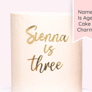 Puede incluir: Un pastel blanco con letras doradas que dicen "Sienna is three".