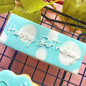 May include: A rectangular cookie with a light blue frosting base and white polka dots. The words "Happy Birthday" are written in white cursive across the top. The cookie is on a black wire rack.