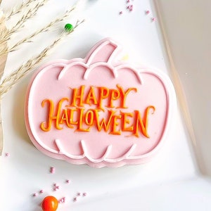 May include: A pink sugar cookie shaped like a pumpkin with the words "Happy Halloween" in orange icing.