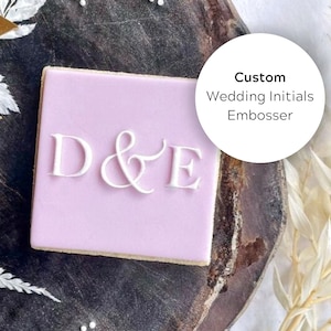 May include: A light pink square cookie with white embossed letters "D & E" on top. The cookie is on a wooden surface with dried flowers and a white circle with the text "Custom Wedding Initials Embosser".