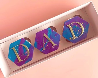 Könnte beinhalten: Drei sechseckige Cupcakes mit lila und blauem Frosting und goldenen Buchstaben, die "DAD" buchstabieren.
