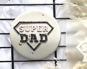 Könnte beinhalten: Ein weißer Kuchen, der mit schwarzem und rosa Zuckerguss in Form eines Diamanten verziert ist, mit den Worten "SUPER DAD" darin.