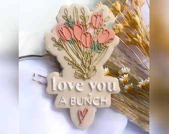 May include: A pink and white cookie decorated with a bouquet of pink tulips and gold leaves. The cookie has the words "love you a bunch" written on it.