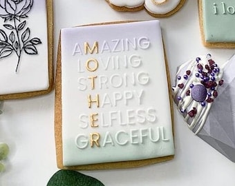 May include: A white and purple decorated cookie with gold lettering spelling out "AMAZING LOVING STRONG HAPPY SELFLESS GRACEFUL" to form the word "MOTHER".