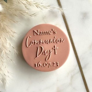 Puede incluir: Un topper de pastel redondo de color rosa con el texto "Name's Communion Day" y un pequeño símbolo de cruz. La fecha "16.07.23" está impresa debajo del texto.