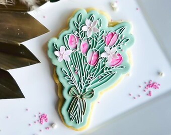 May include: A decorated sugar cookie shaped like a bouquet of flowers. The cookie features pink tulips and white flowers with green leaves, all outlined in white icing on a light green background. A gold bow ties the stems together. Pink sprinkles are scattered around the cookie.