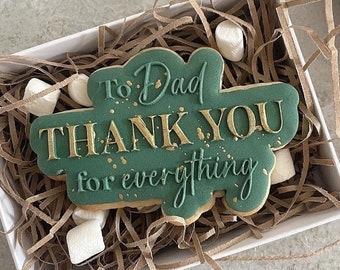 May include: A gift box with a green cookie that says "To Dad THANK YOU for everything" in gold lettering. The cookie is surrounded by brown paper and marshmallows. A spool of twine is visible.