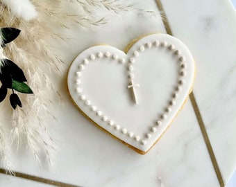 May include: White heart-shaped cookie decorated with a white icing design of a rosary and cross.