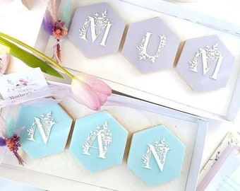 May include: A gift box filled with six hexagonal cookies decorated with white icing and floral designs. The cookies spell out "MUM" and "NAN" in white icing. The box is lined with white paper and has a white ribbon tied around it.