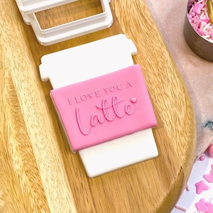 May include: A pink and white cookie cutter in the shape of a coffee cup with the text "I love you a latte" on the pink portion of the cutter.