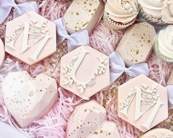 May include: A pink and white gift box filled with decorated cookies. The cookies are shaped like hearts, hexagons, and a cupcake. The cookies are decorated with gold glitter, white icing, and floral designs. The box is filled with pink tissue paper.