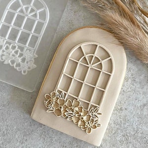 May include: A clear plastic cookie cutter with a design of an arched window with a floral garland. A decorated cookie with a similar design is shown next to the cutter.