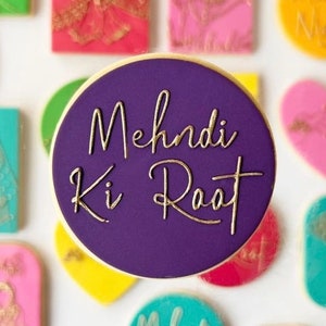 May include: A circular purple cookie with the gold script text "Mehndi Ki Raat". The cookie is surrounded by other colourful cookies in various shapes and colours, including pink, green, and yellow.