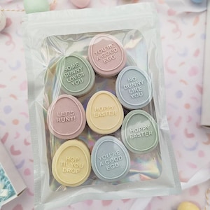 Puede incluir: Una bolsa de plástico transparente llena de huevos de chocolate de color pastel. Cada huevo tiene un mensaje diferente con temática de Pascua, como "Eres un buen huevo", "Feliz Pascua" y "¡Vamos a cazar!"