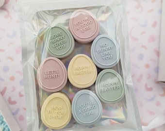 Op de afbeelding: Een doorzichtige plastic zak gevuld met pastelkleurige chocolade-eieren. Elk ei heeft een andere boodschap met een paasthema, zoals "Je bent een goed ei", "Prettige Pasen" en "Laten we jagen!"
