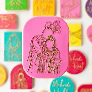 May include: Pink cookie with a gold outline of a couple in traditional Indian wedding attire. The cookie is decorated with intricate details and is ideal for a wedding celebration.