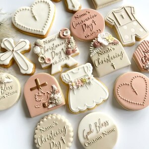 Puede incluir: Surtido de galletas de az&uacute;car decoradas con varios dise&ntilde;os, incluyendo una cruz, un coraz&oacute;n, un vestido, una paloma y un mameluco para beb&eacute;. Las galletas est&aacute;n decoradas con glaseado rosa, blanco y dorado y presentan el texto "Name's Christening Day" y "God Bless Custom Name".